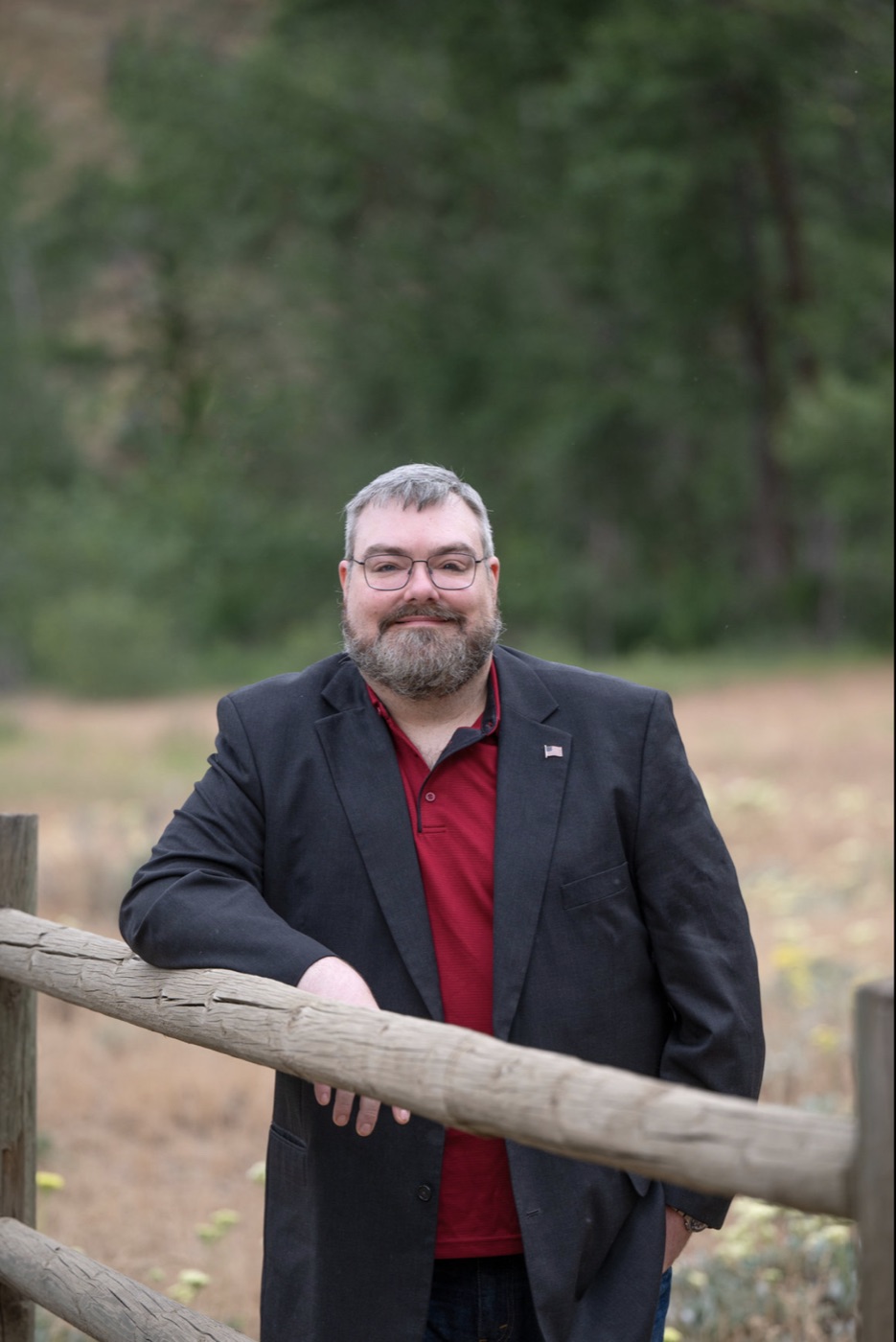 Joshua Thompson, bearded and wearing glasses with a dark blazer and American flag lapel pin, leaning on a wooden rail fence in Central Washington